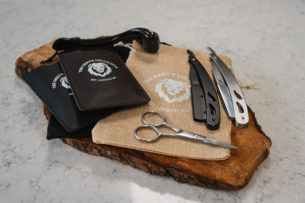Beard Comb Wallets, Black & Brown Drawstring Bags, Black & Silver Straight Razors, Black Derma Roller and Silver Scissors all on a wooden placemat